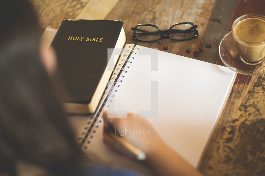 a woman writing in a journal sitting at a table with a Bible 