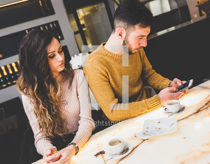 Young couple at the bar. He uses the phone and she is worried and sad. Concept of betrayal.