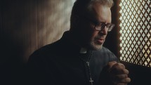 Priest holding hands clasped and saying prayer of absolution in confession booth, penitent sitting behind perforated screen
