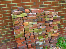 A pile of red stacked loose bricks stacked against a red brick wall outdoors waiting  to be used in construction of a new sidewalk or addition.  