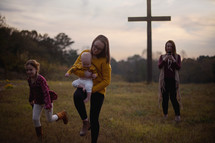 a mother taking pictures of her daughters running in a meadow near a cross