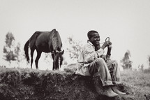 Smiling boy sitting on dirt mound in field with trees holding rope tied to horse.