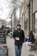 Bearded man walking on the sidewalk near an outdoor coffee cafe.