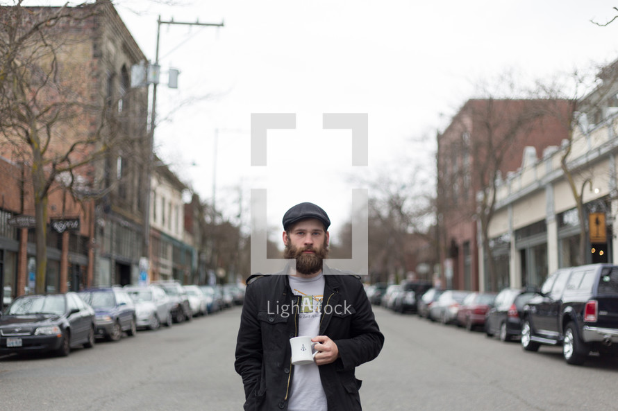 Bearded man with a cup of coffee standing in the middle of the street in town.