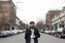 Bearded man with a cup of coffee standing in the middle of the street in town.