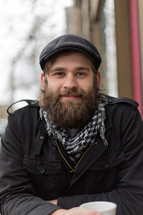 Bearded man with coffee sitting outside smiling.