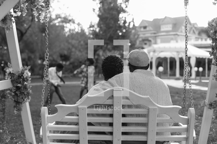 mother and father sitting on a swing watching their children play 