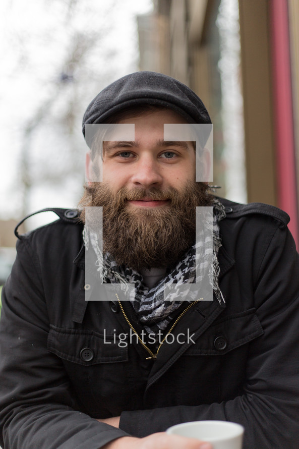 Bearded man with coffee sitting outside smiling.