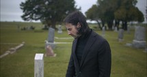 Young, sad man in black suit in cemetery at graveyard tombstone grieving in cinematic slow motion.