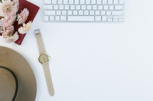 computer keyboard, watch, hat, flowers, and journal on a white desk 
