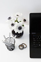 flowers in a black vase, laptop computer, journal, and pens on a desk 