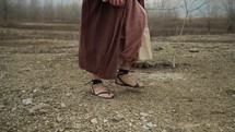 The feet of Jesus Christ dressed in sandals,  brown robes and tunic walking alone in cinematic, slow motion in the wilderness temptation by the Devil or Satan for 40 days and 40 nights fasting and praying. Could also be used to depict a biblical prophet like Noah, Abraham, Elijah, Moses or John the baptist walking and traveling.