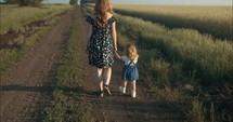 Mother and daughter walking on dirt road while holding hands in summer sunset.