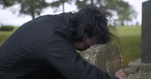 Sad, grieving man embracing family tombstone in cemetery crying in cinematic slow motion. 