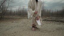 Jesus Christ in the wilderness nature of Jerusalem stoops down to pick up stones and earth from the ground wearing a white tunic and sandals.