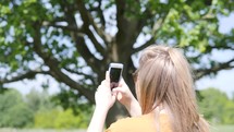 a girl with a phone taking a picture of a tree 