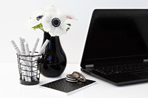 flowers in a black vase, laptop computer, pens, and jar on a desk 