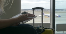 Female traveler using touch pad sitting by the window at airport, suitcse standing nearby. Takeoff strip with planes in background. One airplane is getting off