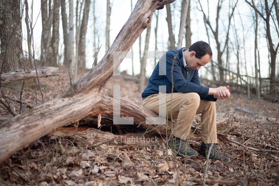 Man sitting outside and praying with folded hands and bowed head in the forest on a fallen tree log
