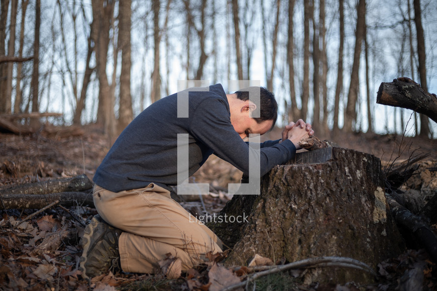 Man kneeling to pray with folded hands on tree stump in the forest