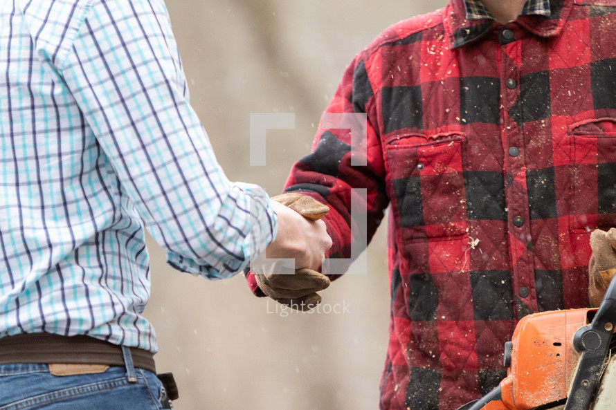White collar man in formal attire shaking hands with blue collar working outdoorsman holding chainsaw outside