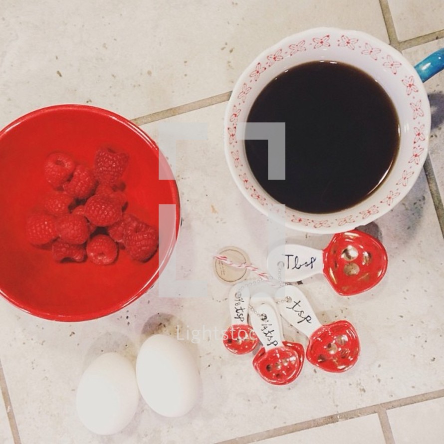 Ingredients on tile: coffee, raspberries, eggs, and measuring spoons.