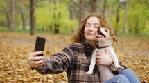 Smiling young woman with dog outdoors in autumn making selfie.