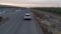 Drone footage of white car on long, empty road near orchards
