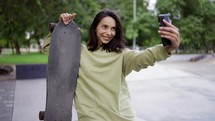 Portrait of a brunette in a green sweater, who sits on the background of the park with a skateboard in her hands and takes a selfie. Walk in the park, hobby, selfie, leisure.