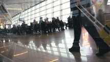 Steadicam low angle shot of a woman walking along the crowded airport lounge with trolley bag. View through the glass