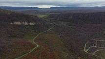 The drone is moving along a winding road between hills, winding through a dense forest