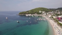 Aerial of tranquil waters, boats, white-sand beach, by picturesque Himare village on Ionian Sea coastline.