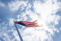 Upward view of sun shining on American flag pole with blue sky and clouds in background