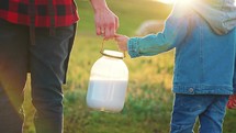 Hands father and son hold can fresh cow milk from farm against an agricultural area and grazing cow in meadow. Natural dairy products. Milking livestock in rural village. Parent walking with child.