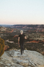 a young man with a backpack standing at the edge of a cliff with a view of canyons 