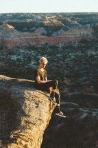 a man sitting a the edge of a cliff at sunset with a view of canyons 