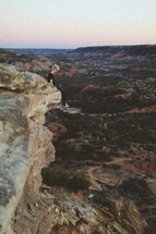 a young man sitting on the edge of a cliff with a view of canyons 