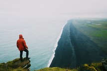 A man standing on a cliff edge above the ocean at a black sand beach by the Dyrhólaey lighthouse in Iceland