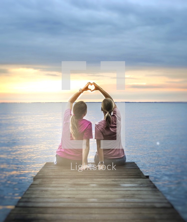 girls sitting at the end of a dock making heart shape with their hands 