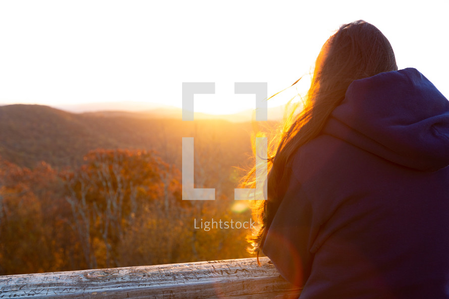 Woman with long hair facing sunlight at overlook enjoying sunrise over autumn trees 