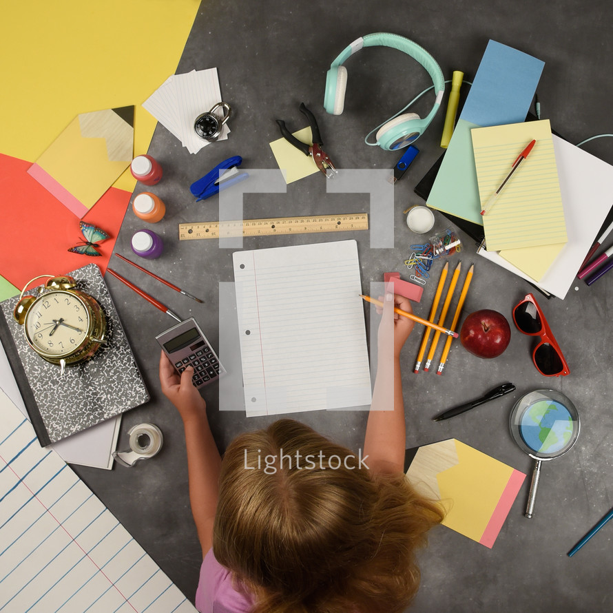 A child is holding a pencil writing on a piece of paper for a back to school education idea. There are various school supplies around the girl.