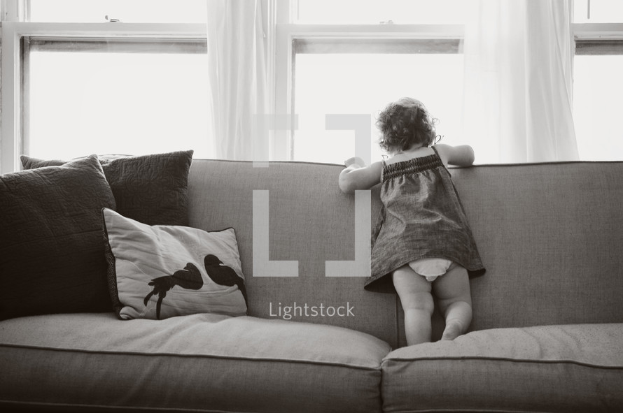 Toddler child standing on a sofa looking out a window.