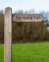 A wood trail sign points the way of the "Permissive Path" with trees and a meadow in the background
