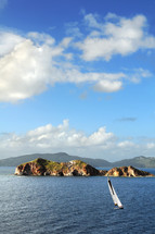 Rock islands in a ocean, with a sailboat in the water, mountains in the background