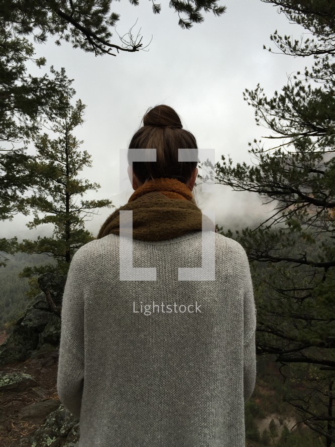 a woman in a sweater with her back to the camera looking out at the foggy valley below 