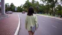 A happy brunette in a green sweater walks with a skateboard in the park. Back view. Walk in the park, portrait. Hobby, leisure.