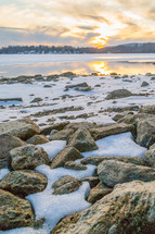 snow on rocks along a shore at sunset 