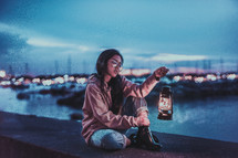 a woman sitting on a concrete wall holding a lantern by a marina 