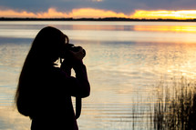 a woman talking a picture of a lake at sunset 