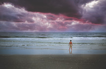 a boy standing on a beach under purple clouds in the sky
